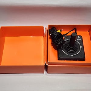 Könnte beinhalten: Ein schwarzer Atari-Joystick-Controller mit einem einzelnen roten Knopf und einem aufgewickelten schwarzen Kabel mit gelbem Stecker, untergebracht in einer leuchtend orangefarbenen Pappschachtel mit passendem Deckel daneben.
