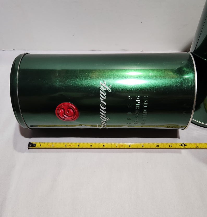以下が含まれることがあります： 白い筆記体で「Tanqueray」の文字が入った、濃い緑色の金属製の円筒形容器。側面に赤いエンブレムがあります。容器の横には巻尺があります。