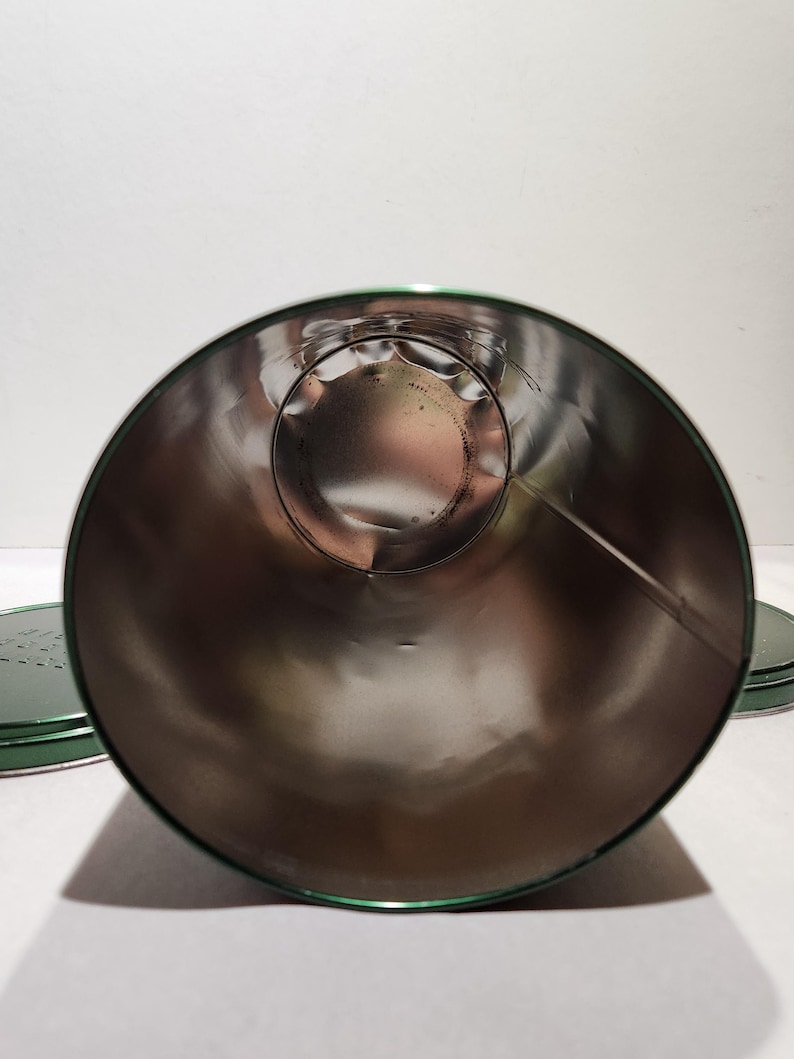 以下が含まれることがあります： 緑色の縁と蓋が付いた、開いた円筒形の金属容器。内側は光沢のある銀色です。容器は空で、スズまたは同様の金属でできているようです。蓋は背景に見えます。
