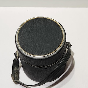 May include: A black, cylindrical case with a silver-colored rim and a zipper closure. The case has a textured surface and a black strap attached. The case is likely designed to hold a camera lens or other small item.