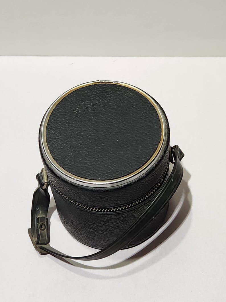 May include: A black, cylindrical case with a silver-colored rim and a zipper closure. The case has a textured surface and a black strap attached. The case is likely designed to hold a camera lens or other small item.
