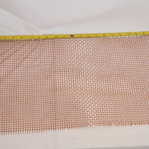 May include: A copper-colored metal mesh screen with a yellow measuring tape showing inches. The mesh is rectangular and has a grid pattern. Several coins are visible near the tape measure.
