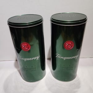 Op de afbeelding: Twee groene metalen Tanqueray gin-blikken met een rood embleem en de merknaam in witte letters. De blikken hebben een cilindrische vorm met een deksel en de woorden "CRISP, REFRESHING, DISTINCTIVE" erop.