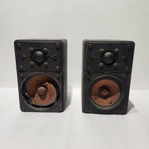 Pode incluir: Dois alto-falantes pretos vintage com cones de alto-falante cor de cobre expostos. Cada alto-falante tem formato quadrado com parafusos visíveis e um elemento central arredondado. Os alto-falantes estão colocados em uma superfície branca.