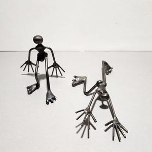 Può includere: Due sculture metalliche di omini stilizzati. Una figura è in piedi con le braccia e le gambe tese. L'altra figura è sdraiata con le braccia e le gambe tese. Entrambe le figure hanno una finitura metallica scura.