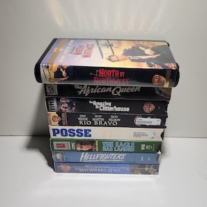 May include: A vertical stack of eight VHS tapes featuring titles including North by Northwest, The African Queen, The Amazing Dr. Clitterhouse, Rio Bravo, Posse, The Eagle Has Landed, Hellfighters, and The Man Without a Face with various spine designs.