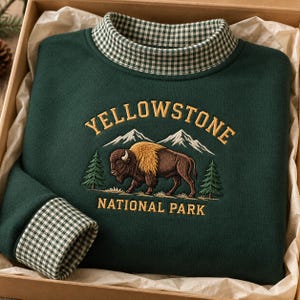 May include: A dark green sweatshirt featuring a mock neck collar with a green and white gingham pattern. The chest displays an embroidered graphic of a brown bison, two pine trees, and snow-capped mountains with the text YELLOWSTONE NATIONAL PARK.