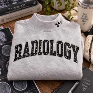 May include: A light gray mock neck sweatshirt featuring the word RADIOLOGY in black block letters with white polka dots across the chest. A small black radiation symbol and a heart are embroidered on the right side of the neckline.
