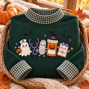 May include: A dark green long-sleeve sweatshirt with a mock neck and cuffs featuring a green and white gingham pattern. The front displays an embroidered graphic of a ghost, stethoscope, mummy, jack-o'-lantern pill bottle, and clipboard.