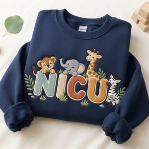May include: A navy blue crewneck sweatshirt featuring embroidered text that reads NICU. The letters are decorated with a lion, an elephant, a giraffe, and a zebra, surrounded by small green leaves and grass accents on the front of the garment.