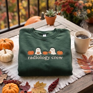 May include: A dark green crewneck sweatshirt featuring an embroidered design of two white ghosts and three orange pumpkins. The text "radiology crew" is embroidered in white below the ghosts and pumpkins. The sweatshirt is folded on a white cloth.