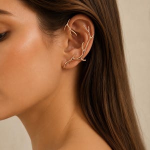 Puede incluir: Un pendiente tipo ear cuff de tono dorado con forma de cornamenta ramificada que se envuelve alrededor del borde exterior de la oreja. La pieza presenta un acabado metálico liso con múltiples puntas que se extienden por el cartílago y el lóbulo.