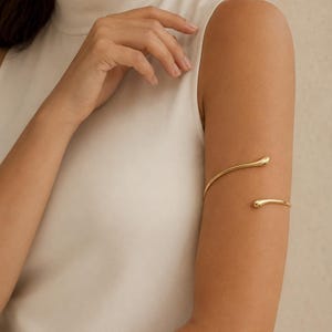 Op de afbeelding: Een goudkleurige metalen armband voor de bovenarm met een strak, open ontwerp en twee afgeronde, druppelvormige uiteinden. De gepolijste, gladde band wikkelt zich gedeeltelijk om de bovenarm voor een doorlopend metalen silhouet.