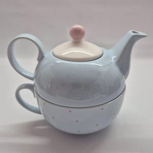 May include: A light blue ceramic tea set for one, featuring a teapot stacked on a matching teacup. The teapot has a white lid with a small pink knob. The teacup is decorated with small pink polka dots and has a curved handle on the side.