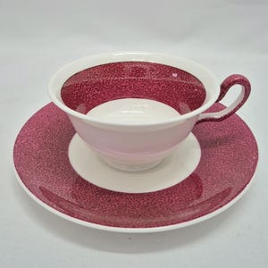 May include: A ceramic teacup and saucer set featuring a white base with a deep magenta speckled pattern. The teacup has a curved handle and a wide rim, while the saucer has a matching wide magenta border. The interior of the cup is white.