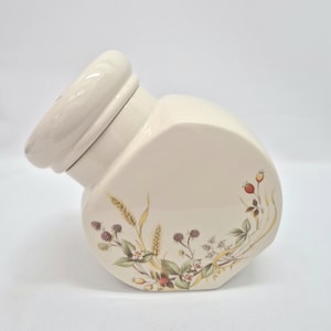 Peut inclure: Un pot en céramique de couleur crème avec un couvercle assorti, orné d'une illustration botanique de mûres, d'épis de blé et de petites baies rouges. Le récipient présente une forme asymétrique angulaire unique avec une finition lisse et brillante.