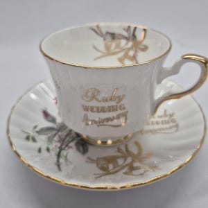 Może przedstawiać: Biały porcelanowy zestaw filiżanki ze spodkiem ze złotym wykończeniem. Filiżanka posiada złoty napis "Ruby Wedding Anniversary" oraz wzór kokardy. Spodek ozdobiony jest motywem kwiatowym z zielonymi liśćmi, różowymi pąkami i złotymi kokardami.