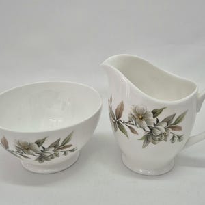 May include: A white porcelain creamer and sugar bowl set featuring a delicate floral pattern with muted green and brown leaves and blossoms. Both pieces have a smooth, rounded shape with a pedestal base and a glossy finish on the exterior.