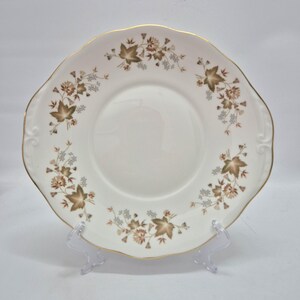 May include: A round white ceramic plate with a scalloped edge and a gold-trimmed rim. The plate features a repeating pattern of brown maple-like leaves and small light blue floral clusters arranged in a circular design around the center.
