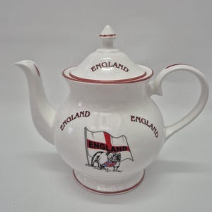Puede incluir: Una tetera de cerámica blanca con borde rojo, asa curva y pico. La palabra ENGLAND está impresa en rojo en la tapa y el cuerpo. En la parte frontal hay un gráfico de un bulldog frente a una bandera ondeante con la palabra ENGLAND.