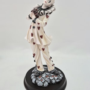 Könnte beinhalten: Eine Clown-Figur mit blasser Haut und dunklem Augen-Make-up, bekleidet mit einem cremefarbenen Outfit, das mit kleinen roten herzförmigen Aufnähern und einem schwarzen Rüschenkragen verziert ist. Der Clown hält dunkle Rosen auf einem Steinsockel.