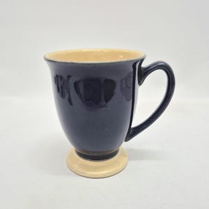 May include: A ceramic mug with a glossy dark navy blue exterior and a smooth, light cream-colored interior. The mug features a flared rim, a curved handle on the right side, and a distinct, raised circular base in a matching light cream color.