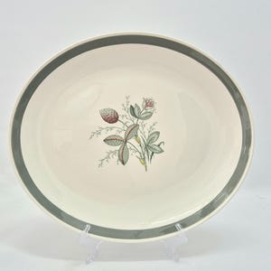 Può includere: Un piatto da portata ovale in ceramica color bianco sporco. Il bordo presenta un'ampia fascia solida di colore grigio scuro. Il centro del piatto mostra un'illustrazione botanica dettagliata di una pianta di trifoglio con foglie verdi e boccioli.