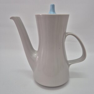 Puede incluir: Una cafetera de cerámica gris claro con un cuerpo alto y esbelto, un asa curva y un pico largo e inclinado. La tapa presenta un pequeño pomo cónico de color azul claro centrado en la parte superior. La superficie es lisa y mate.