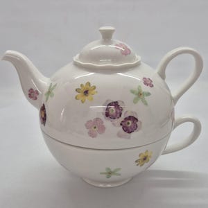 Puede incluir: Un juego de té individual de cerámica blanca que consta de una tetera con tapa y una taza a juego apilada debajo. El conjunto presenta un delicado patrón de pequeñas flores prensadas en tonos amarillo, rosa, morado y verde claro.