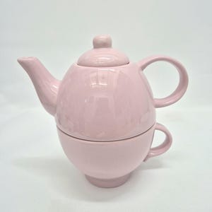 Puede incluir: Un juego de té individual de cerámica en color rosa claro, con un diseño apilable en el que una tetera descansa sobre una taza a juego. La tetera tiene un pico curvo, una tapa redondeada con un pequeño pomo y un asa, mientras que la taza cuenta con su propia asa.