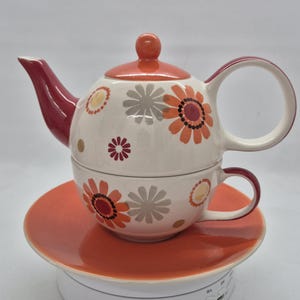 Op de afbeelding: Een keramische theeset voor één persoon bestaande uit een theepot gestapeld op een theekopje, rustend op een bijpassende schotel. De set is versierd met een herhalend patroon van oranje, rode en grijze gestileerde bloemen op een crèmekleurige basis.