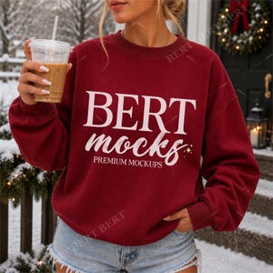 以下が含まれることがあります： 深い赤色のクルーネックのスウェットシャツで、白い文字で「BERT mocks PREMIUM MOCKUPS」と小さな星のアイコンがプリントされています。長袖でリブ編みの襟元があり、水色のデニムショートパンツと組み合わされ、透明なプラスチックカップを持っています。