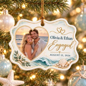 May include: A white, scalloped-edge ceramic ornament featuring a heart-shaped photo of a couple on a beach. The text reads Olivia & Ethan, Engaged, and AUGUST 15, 2026. The design includes watercolor ocean waves, green leaves, pearls, and seashells.