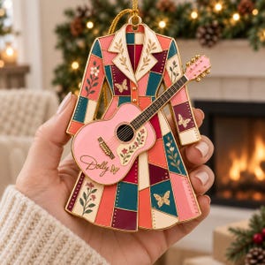 May include: A gold-toned metal ornament shaped like a long coat with a patchwork design in shades of pink, teal, and burgundy. The coat features floral and butterfly motifs and has a pink acoustic guitar attached to the front with the name Dolly.