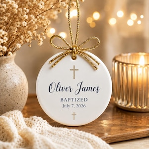 May include: A round white ceramic ornament featuring a gold cross at the top and bottom. The text reads Oliver James, BAPTIZED, and July 7, 2026, in dark blue lettering. A gold metallic cord is tied in a bow at the top for hanging purposes.