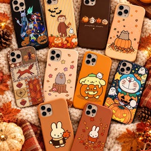 May include: A collection of ten smartphone cases featuring various autumn-themed illustrations, including characters like Pusheen, Miffy, and Doraemon, alongside pumpkins, fall leaves, and seasonal motifs, all arranged on a textured cream-colored surface.