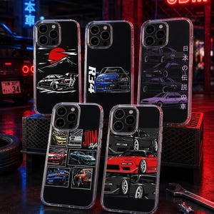 May include: Five clear phone cases featuring various Japanese sports car illustrations. Designs include a red sun with a mountain, a blue R34 model with text, a column of cars with Japanese text, a grid of six car images, and a red car graphic.