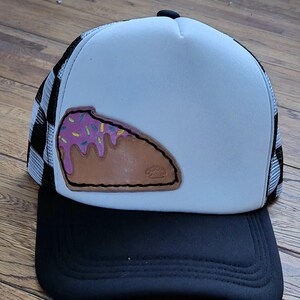May include: A white and black trucker hat featuring a tan leather patch shaped like a slice of cake with pink frosting and colorful sprinkles. The hat has a solid black brim, a white front panel, and a black and white checkered mesh back panel.