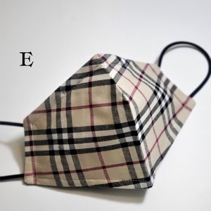 May include: A beige, black, and red plaid face mask with black elastic ear loops. The mask has a 3D shape and is made of fabric. The letter "E" is visible in the top left corner of the image.