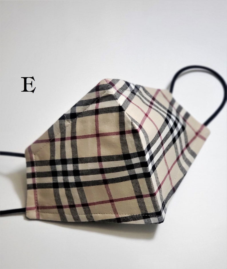 May include: A beige, black, and red plaid face mask with black elastic ear loops. The mask has a 3D shape and is made of fabric. The letter "E" is visible in the top left corner of the image.