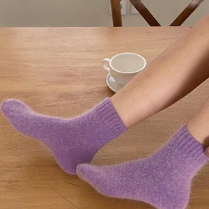 May include: A pair of ankle-length socks in a soft, fuzzy lavender knit material. The socks feature a ribbed cuff at the top and a solid, uniform purple color throughout, with a visible texture that appears plush and warm for everyday wear.