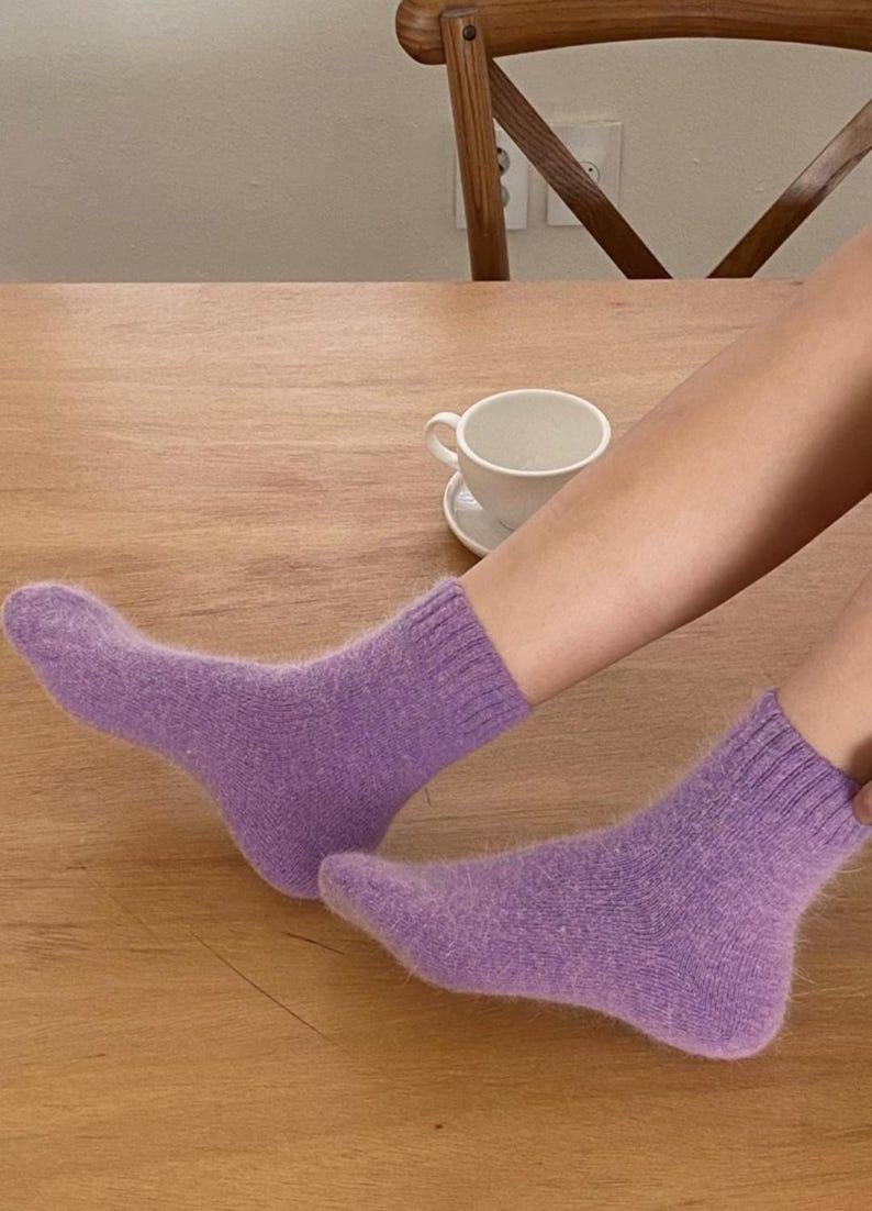May include: A pair of ankle-length socks in a soft, fuzzy lavender knit material. The socks feature a ribbed cuff at the top and a solid, uniform purple color throughout, with a visible texture that appears plush and warm for everyday wear.