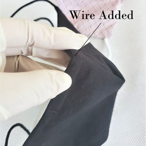 May include: A black face mask with a wire added to the top edge. The wire is visible through the fabric. The mask is being held by a gloved hand.