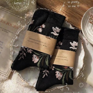 May include: Two pairs of black socks with a floral pattern, wrapped with brown paper bands. The socks feature white and pink flowers with green leaves. The text "breezemountainB.etsy.com" is visible on the bands. The socks are on a white, scalloped plate.