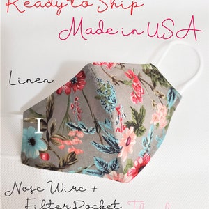 May include: A gray linen face mask with a floral print. The mask has a nose wire and a filter pocket. The text "Ready to Ship Made in USA" is written above the mask. The text "Linen" is written below the mask. The text "Nose Wire + Filter Pocket" is written below the mask. The text "Thank you" is written below the mask.