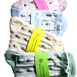 May include: Four pairs of pastel-colored socks are stacked. Each sock has a floral pattern and a ruffled edge. The socks are in shades of pink, blue, cream, and green. Each pair is wrapped with a tag that includes a website address.