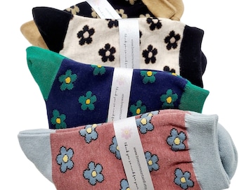 Op de afbeelding: Vier paar sokken met bloempatronen. De sokken hebben verschillende kleurencombinaties, waaronder zwart en beige, marineblauw en groen en roze en grijs. Elk paar is verpakt met een witte band met een websiteadres.