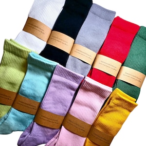 May include: A collection of ten pairs of colorful ribbed socks. The socks are folded and arranged in a fan shape. The socks are all different colors, including green, red, yellow, pink, lavender, light blue, light green, gray, black, and white. Each pair of socks has a brown paper tag with the website address "www.treemouthstudio.etsy.com".