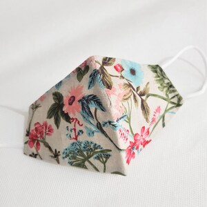 May include: A beige face mask with a floral pattern featuring pink, blue, and green flowers. The mask has white elastic ear loops.