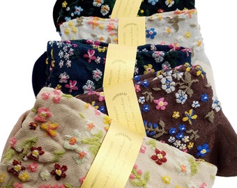 Op de afbeelding: Een stapel sokken met bloemenpatroon in verschillende kleuren, waaronder marineblauw, bruin en beige. Elk paar is verpakt met een gouden lint. De sokken hebben geborduurde bloemen in tinten roze, geel en blauw.